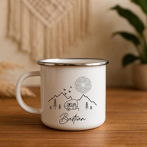 May include: White enamel camping mug with a black mountain and camper design. The mug features a sun, birds, and trees, with the name "Bastian" in script. The mug has a silver rim and handle.