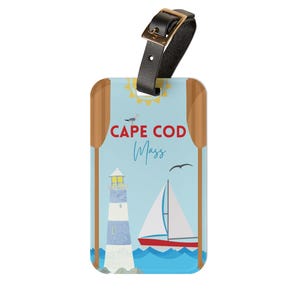 May include: Luggage tag with a Cape Cod, Massachusetts design. The tag features a lighthouse, sailboat, and the text "Cape Cod Mass" on a light blue background. It has a black leather strap with a gold-coloured buckle.