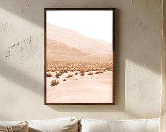 May include: A framed landscape print depicting a desert scene. The artwork features a mountain range in the background, with sand dunes and sparse vegetation in the foreground. The color palette is primarily warm tones of beige and brown.