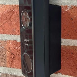 Puede incluir: Timbre de seguridad Eufy negro rectangular montado en una pared de ladrillos rojos. El timbre tiene una lente de cámara, un botón circular y el logotipo de Eufy. La pared de ladrillo tiene una textura rugosa.