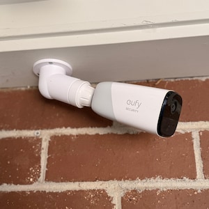 May include: A white and light grey Eufy Security camera mounted on a wall. The device features a cylindrical body with a black front panel containing the lens. The camera is attached to a white adjustable mounting bracket secured to the surface.