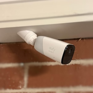 May include: A white and grey Eufy Security outdoor camera mounted to a white horizontal surface. The device features a cylindrical body with a black front face containing the lens and sensor, connected to a white adjustable mounting bracket.