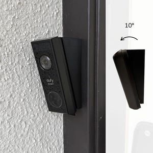 Puede incluir: Timbre de video eufy Security negro montado en una pared texturizada blanca. El timbre tiene una lente de cámara, un botón y el logotipo de eufy. El soporte de montaje también es negro.