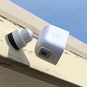 May include: A white Eufy Security camera mounted on a surface, featuring a square-shaped housing with a black circular lens base, a swivel joint, and a QR code label on the bottom panel with a series of small ventilation holes near the edge.