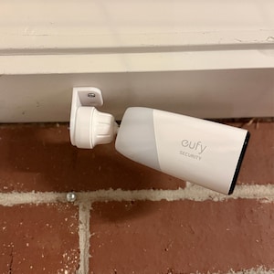 May include: A white Eufy Security camera mounted horizontally to a white surface. The device features a cylindrical body with a black lens face and a swivel mount attached by a single visible screw. The brand name Eufy Security is printed in gray.