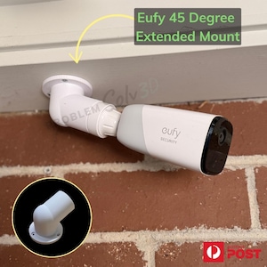 May include: A white Eufy security camera mounted on a 45-degree extended mount. The camera is attached to a white bracket, which is mounted on a brick wall. The text "Eufy 45 Degree Extended Mount" is visible.