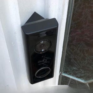 May include: A black Eufy Security video doorbell mounted at an angle on a white door frame. The device features a circular camera lens at the top, the Eufy Security logo in the centre, and a circular doorbell button at the bottom of the unit.