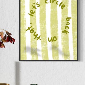 May include: A framed art print with a green and white striped background. The text "let's circle back on that" is written in a circular pattern in green. The artwork is displayed on a white wall above a gold side table.
