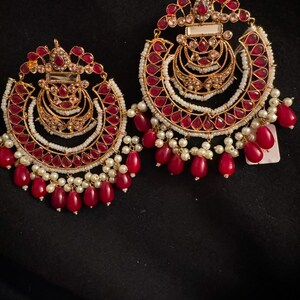 May include: A pair of large, gold-tone earrings with red and white beads. The earrings are circular in shape and have a decorative design with multiple layers of beads. The earrings are accented with small, clear stones.