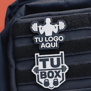 Puede incluir: Dos parches en blanco y negro con diseños de fitness. Uno presenta la silueta de un levantador de pesas y el texto "TU LOGO AQUI". El otro muestra "TU BOX" con pesas y una barra. Ambos sobre un fondo azul oscuro.