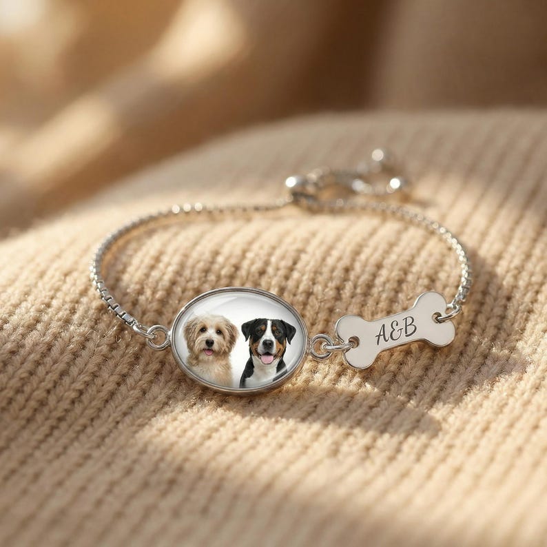 K&ouml;nnte beinhalten: Silbernes Armband mit einem ovalen Anh&auml;nger mit zwei Hundeportr&auml;ts und einem knochenf&ouml;rmigen Charm. Der Knochen-Charm tr&auml;gt die Initialen "A&B". Das Armband wird auf einer beigefarbenen, strukturierten Oberfl&auml;che gezeigt.