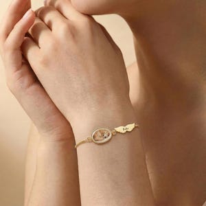 K&ouml;nnte beinhalten: Goldfarbenes Armband mit einem ovalen Anh&auml;nger, der ein Foto eines schlafenden Hundes zeigt. Ein fl&uuml;gelf&ouml;rmiger Charm mit dem Namen "Kiki" ist angebracht. Das Armband wird am Handgelenk getragen.