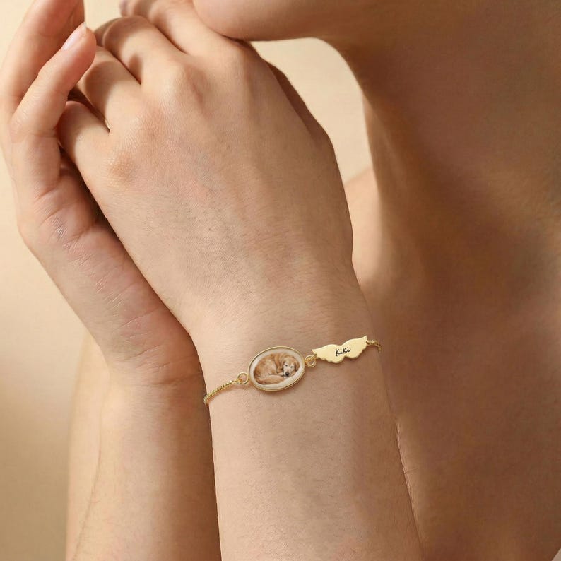 K&ouml;nnte beinhalten: Goldfarbenes Armband mit einem ovalen Anh&auml;nger, der ein Foto eines schlafenden Hundes zeigt. Ein fl&uuml;gelf&ouml;rmiger Charm mit dem Namen "Kiki" ist angebracht. Das Armband wird am Handgelenk getragen.