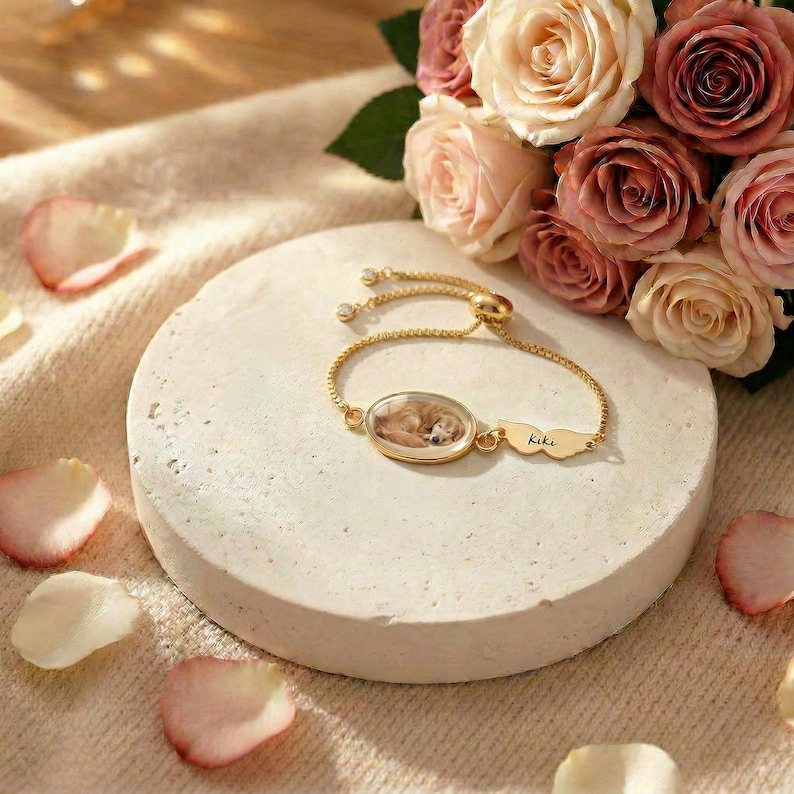 K&ouml;nnte beinhalten: Ein goldfarbenes Armband mit einem ovalen Anh&auml;nger, der ein schlafendes Hundeabbild zeigt. Das Armband hat eine feine Kette und einen fl&uuml;gelf&ouml;rmigen Charm mit dem Namen "Kiki". Im Hintergrund befinden sich ein Rosenstrau&szlig; und verstreute Bl&uuml;tenbl&auml;tter.