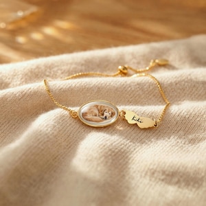 K&ouml;nnte beinhalten: Ein goldfarbenes, verstellbares Armband mit einem ovalen Anh&auml;nger, der ein Portr&auml;t eines schlafenden Hundes zeigt. Das Armband enth&auml;lt einen Charm mit dem eingravierten Namen "Kiki". Die Kette ist filigran und liegt auf beigem Stoff.