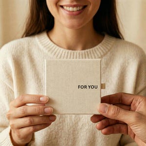 K&ouml;nnte beinhalten: Eine beige Geschenkbox mit dem schwarzen Aufdruck "FOR YOU". Die Box wird von zwei Personen gehalten. Die Person im Hintergrund tr&auml;gt einen cremefarbenen Pullover.