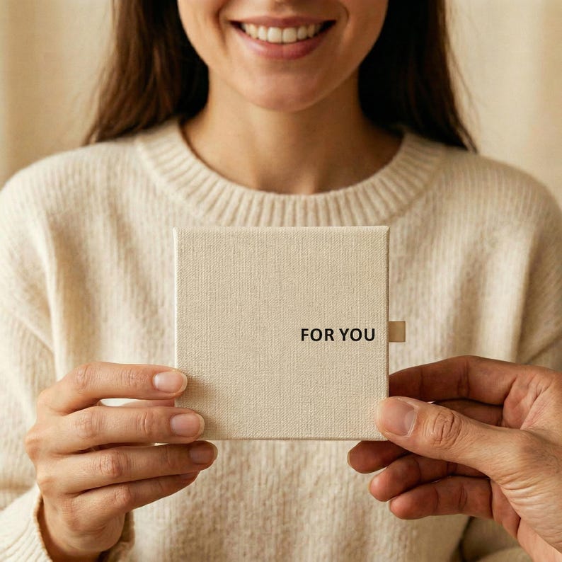 K&ouml;nnte beinhalten: Eine beige Geschenkbox mit dem schwarzen Aufdruck "FOR YOU". Die Box wird von zwei Personen gehalten. Die Person im Hintergrund tr&auml;gt einen cremefarbenen Pullover.