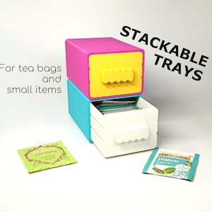 May include: Stackable trays in vibrant colours, ideal for organising tea bags and small items. The trays are pink, yellow, blue, and white. The image also includes two tea bag packets, and the text "For tea bags and small items" and "STACKABLE TRAYS".