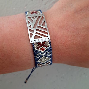 Pode incluir: Um bracelete de contas azuis e prateadas com um amuleto geométrico prateado. O bracelete tem um padrão vermelho e branco.