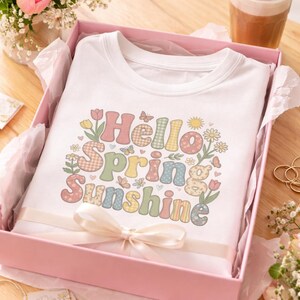Puede incluir: Una camiseta blanca con las palabras "Hello Spring Sunshine" en letras coloridas con temática floral. La camiseta se presenta en una caja de regalo rosa con una cinta blanca, rodeada de flores y una bebida.