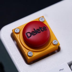 May include: A mechanical keyboard keycap featuring a circular red button with the word Delete printed in black. The button is mounted on a square yellow base secured by four silver-colored screws, positioned on a white keyboard chassis.