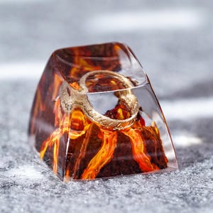 May include: A translucent resin mechanical keyboard keycap featuring a miniature golden ring embedded within a dark brown and orange volcanic landscape design. The keycap has a smooth, glossy finish and a sloped, trapezoidal shape with a clear top.