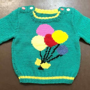 Könnte beinhalten: Ein teal-farbener Strickpullover mit gelbem Ausschnitt und Bündchen. Der Pullover hat ein farbenfrohes Ballondesign in Pink, Gelb, Rot und Blau. Der Pullover hat zwei Knöpfe auf jeder Schulter.