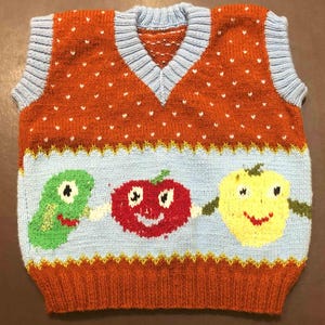 May include: A sleeveless knit sweater vest featuring a rust-orange upper section with small white dots and a light blue lower section. The design includes three cartoon fruit characters in green, red, and yellow, with light blue ribbed trim.