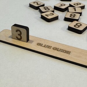 May include: A wooden guide with the text "GLUE GUIDE" printed on it. A small wooden block with the number "3" printed on it is placed on the guide.