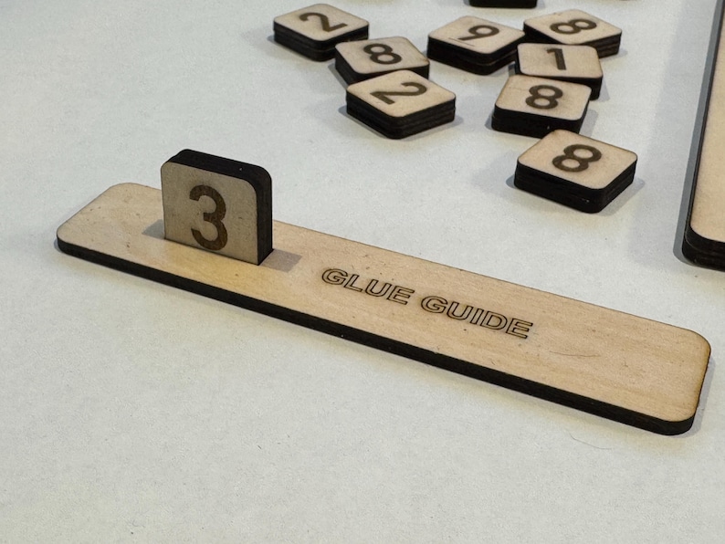 May include: A wooden guide with the text "GLUE GUIDE" printed on it. A small wooden block with the number "3" printed on it is placed on the guide.