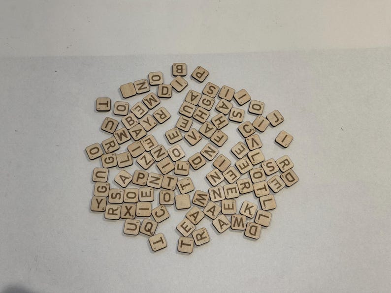 May include: A collection of small, square wooden tiles, each etched with a single letter or number. The tiles are scattered across a white surface, creating a random assortment of characters. The natural wood color provides a neutral backdrop.