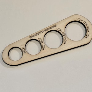 May include: A wooden spaghetti measure with four circular holes of different sizes. The text "SPAGHETTI MEASURE" is engraved on the surface. Each hole has a small engraved figure indicating portion sizes. The wood has a light, natural finish.