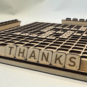 May include: Wooden Scrabble game board with the word "THANKS" displayed on the tile rack. The board is made of light-colored wood with a grid of squares. Other letter tiles are placed on the board. A fun board game.