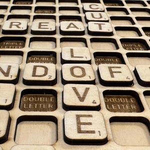 May include: Wooden Scrabble tile set spelling out "CUT GREAT LOVE" with individual letter values. The tiles are set in a wooden grid, with spaces for additional tiles. The letters are in a dark brown color.