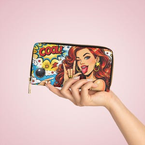 May include: A zip-around wallet with a pop art design of a woman winking and sticking out her tongue. The word "COOL!" is displayed in a comic book style. The wallet has a black zip and a gold-coloured pull.