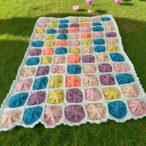 May include: A colorful, handmade crochet blanket with a grid pattern. The blanket features squares of various pastel colors, including pink, blue, yellow, and purple, set against a white background. The blanket is placed on green grass.
