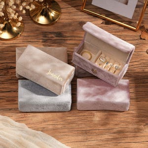 May include: Several velvet ring boxes in shades of beige, grey, and pink. One box is open, revealing rings. The word "Jennifer" is embroidered on a box. The boxes are displayed on a wooden surface.