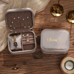 May include: A gray velvet jewelry box, open to reveal a watch, rings, and necklaces. The closed box next to it has the name "Olivia" in gold script. The box is on a wooden surface, with decorative items in the background.