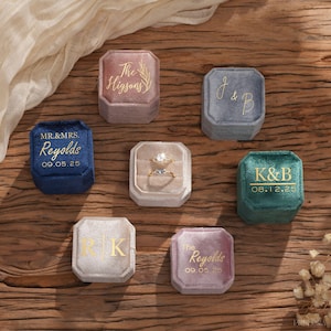 Pode incluir: Uma coleção de caixas de anel de veludo em várias cores, incluindo azul, rosa e verde, com letras douradas. Algumas caixas exibem iniciais, nomes e datas. Uma caixa contém dois anéis. Dispostas sobre uma superfície de madeira.