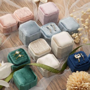Pode incluir: Uma coleção de caixas de anel de veludo em várias cores, incluindo rosa, azul, verde e creme. Algumas caixas estão abertas, exibindo anéis com bandas de ouro e pedras preciosas. As caixas estão dispostas em uma superfície de madeira com detalhes florais.