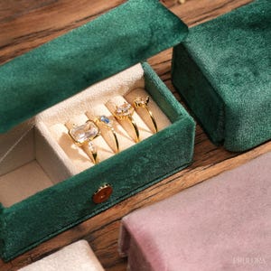 Puede incluir: Una caja de joyería de terciopelo verde con cuatro anillos de oro con piedras preciosas. Los anillos se exhiben en un interior color crema. La caja está abierta, revelando los anillos, y se encuentra sobre una superficie de madera.
