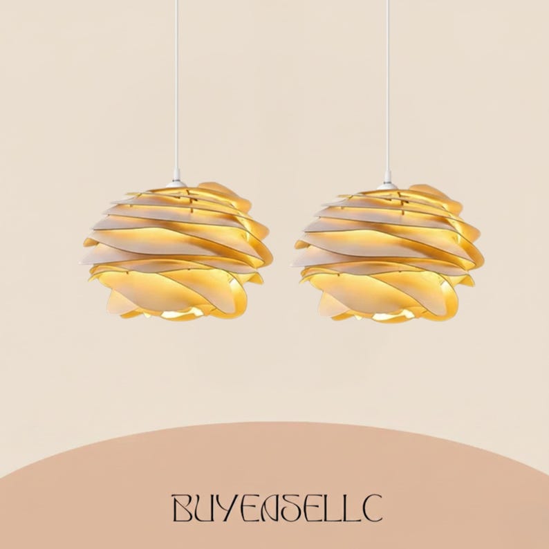 May include: Two pendant lights with a layered, petal-like design. The lights are a warm gold color, with a white interior, and hang from white cords. The lights are suspended against a neutral background. The text "BUYENSELLC" is at the bottom.