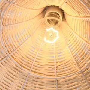 May include: Close-up of a woven rattan lampshade with a white metal frame. The light bulb is visible at the center, emitting a warm glow. The lampshade's design features a natural, light brown color, creating a cozy and inviting atmosphere.