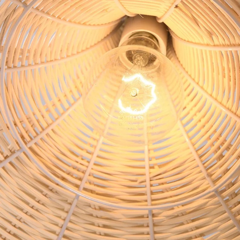 May include: Close-up of a woven rattan lampshade with a white metal frame. The light bulb is visible at the center, emitting a warm glow. The lampshade's design features a natural, light brown color, creating a cozy and inviting atmosphere.