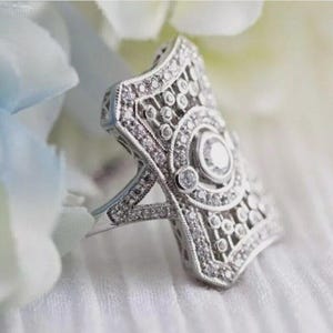 May include: A silver-toned ring with an Art Deco design. The ring features a rectangular face with a central, round, clear stone and is encrusted with smaller, clear stones. The ring's intricate filigree design adds to its vintage appeal.