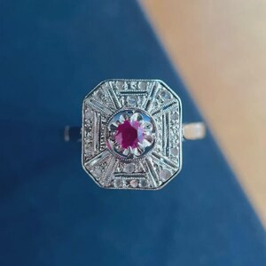 May include: A silver ring with an octagonal face, featuring a central ruby surrounded by smaller clear stones. The ring has intricate filigree detailing and a polished finish, set against a blue background.