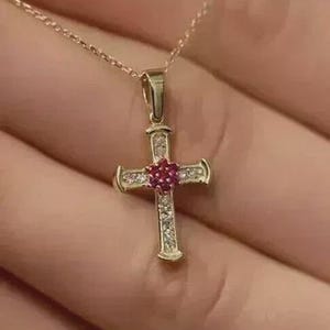 Pode incluir: Um colar com pingente de cruz dourada. A cruz apresenta uma pedra preciosa vermelha central e é adornada com pedras preciosas claras menores. A corrente do colar é delicada e dourada, criando uma joia clássica e elegante.