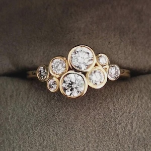 Puede incluir: Un anillo de oro con un grupo de piedras preciosas redondas y transparentes. Cada piedra preciosa está engastada en su propio bisel, creando un diseño distintivo y natural. La banda del anillo es delgada y pulida.