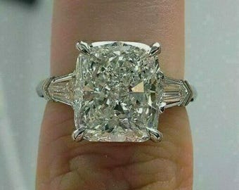 May include: A large, square-cut diamond ring with a platinum band. The ring features a central, cushion-cut diamond flanked by two smaller, baguette-cut diamonds. The ring is set on a finger.