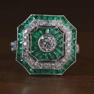 May include: A striking emerald and diamond ring. The ring has a geometric design, with a central clear diamond encircled by emerald-cut emeralds and smaller diamonds. The setting is silver-toned. The ring is displayed on a dark wooden surface.